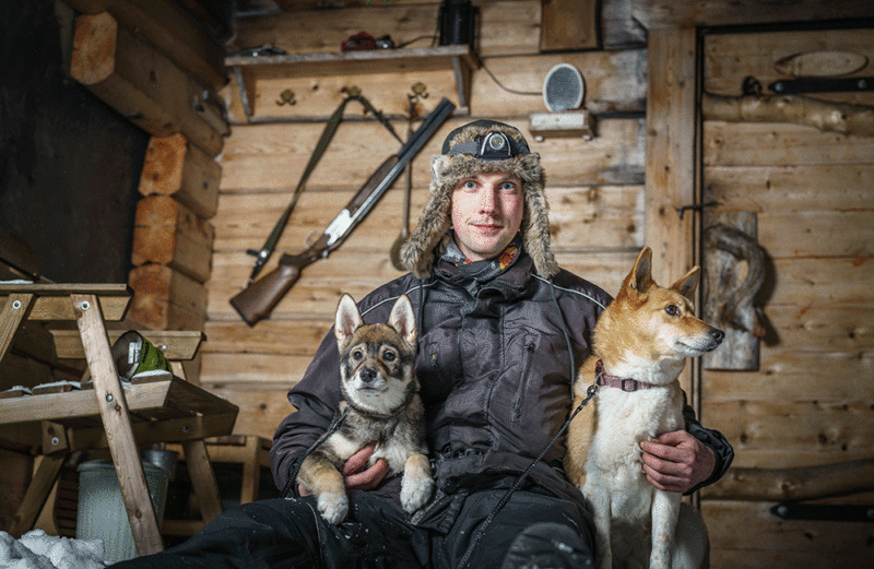 Nuori mies poseeraa metsästysmajassa kahden koiran kanssa.