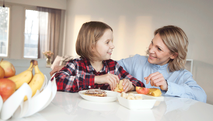 Kouluikäinen tyttö istuu äitinsä kanssa välipalapöydässä.