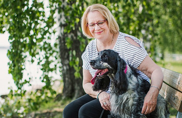 Yli keski-ikäinen vaaleatukkainen nainen istuu puiston penkillä silittäen koiraansa.