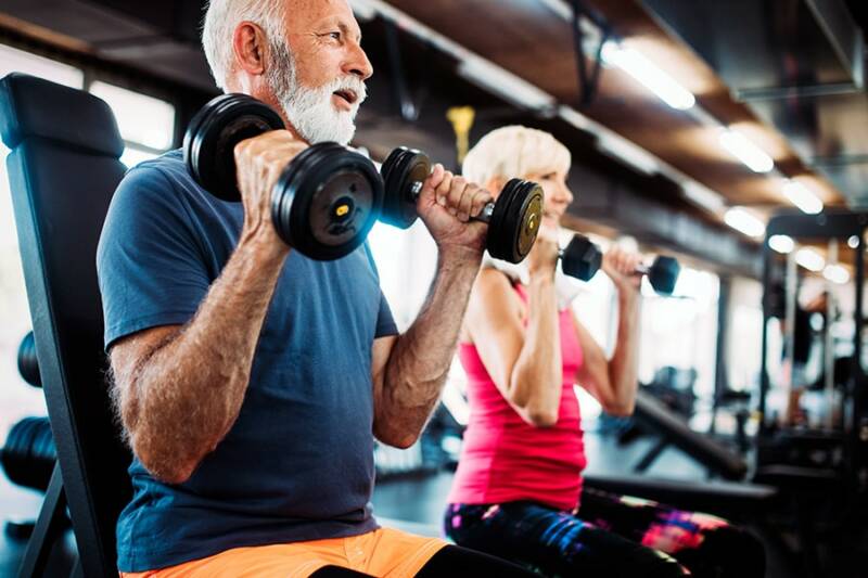 En äldre man lyfter vikter på gymmet med en instruktör bredvid sig.