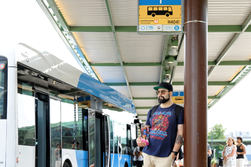 Tomi Heiskanen nojaa pylvääseen bussipysäkillä, hänellä on kädessään Barbie-nukkepaketti